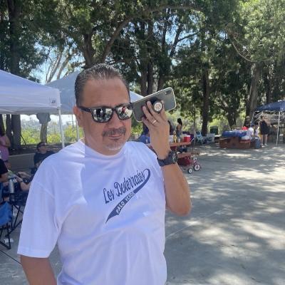 Picture of Eddie Santillan listening to something on his phone. He wears sunglasses and a Los Desterrados tshirt.